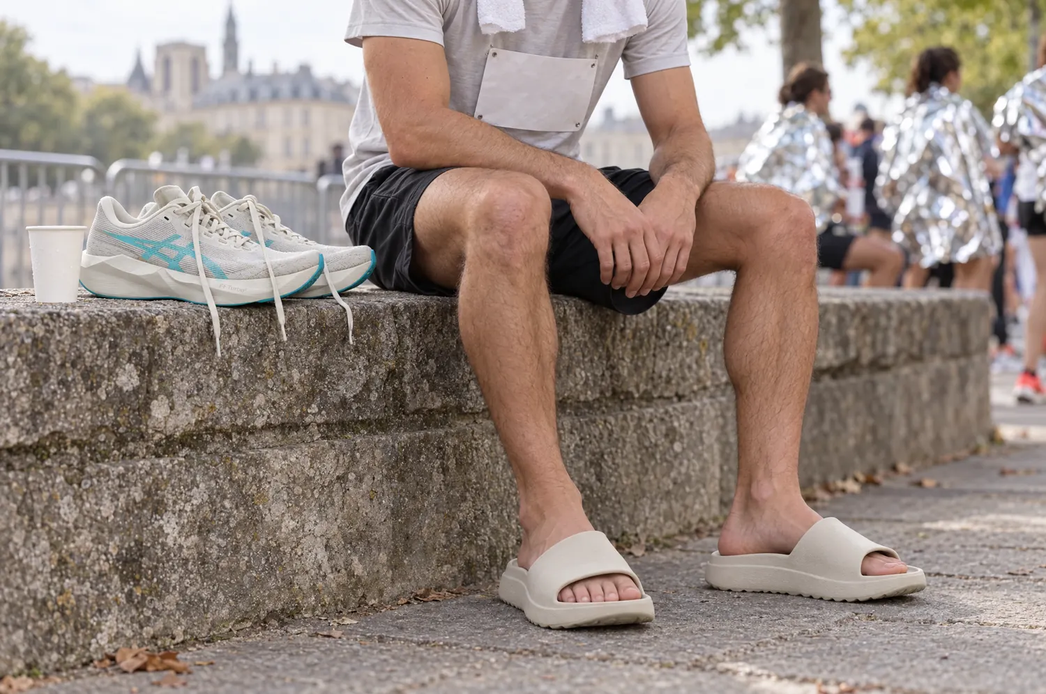 Coureur assis sur un muret après une course sur route, en claquettes de récupération, ses chaussures posées à côté de lui